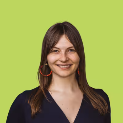 Smiling woman with straight brown hair wearing red hoop earrings and a dark top against a light green background.